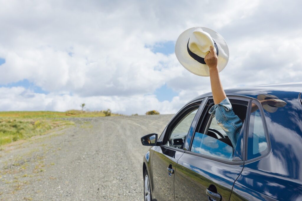 mulher ergue seu chapéu para o céu pela janela do banco de trás do carro em uma estrada deserta