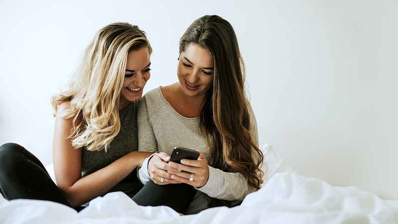 duas amigas conversam sentadas na cama enquanto olham uma conversa no celular