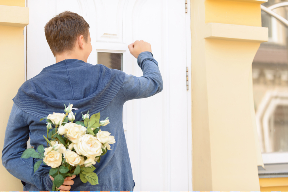 Homem batendo na porta com um buquê de flores atrás da outra mão