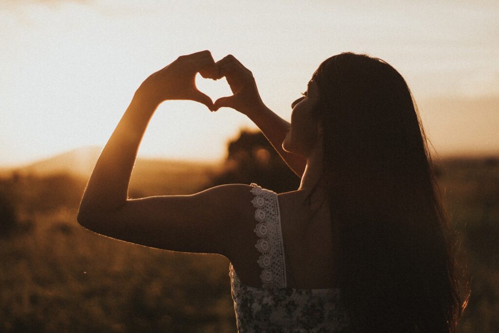 mulher faz coração com as mãos durante o pôr do sol no campo