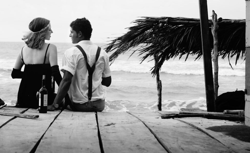 casal sentado em deque na praia com roupas vintage e garrafa de vinho