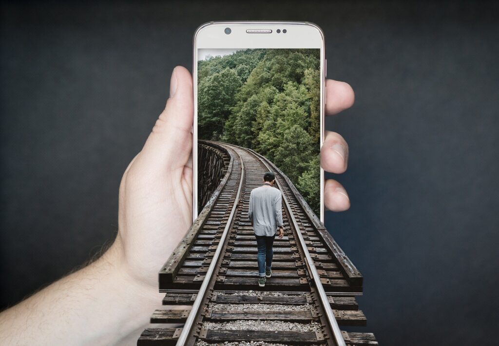 imagem com edição gráfica: trilhos de trem adentram a tela de celular enquanto homem caminha sobre eles e mulher segura o aparelho de celular
