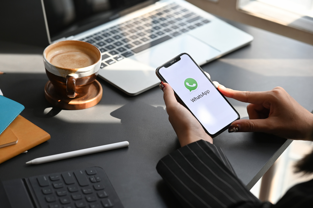 mulher segura o celular com o Whatsapp iniciando em cima de uma mesa com laptop e café