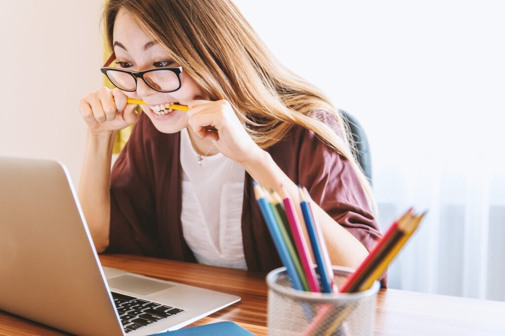 mulher jovem estuda em frente ao computador, com um lápis entre os dentes e expressão alegre