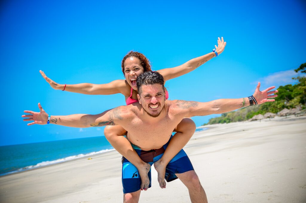 mulher sobre as costas do companheiro, ambos de braços abertos e sorrindo na praia