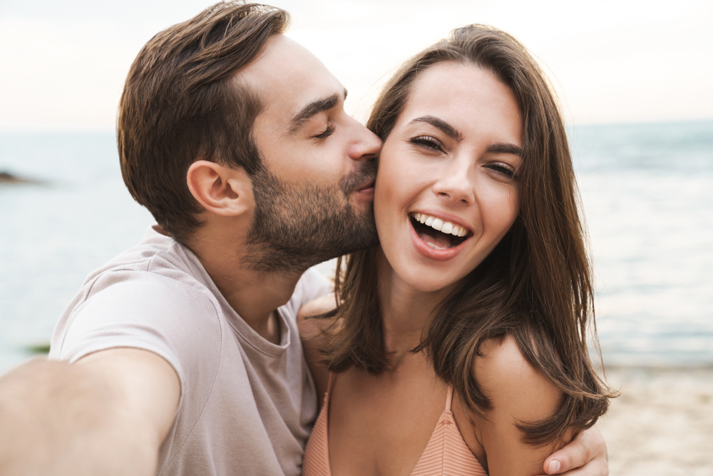 homem beija a namorada no rosto enquanto tiram selfie na praia, a mulher sorri