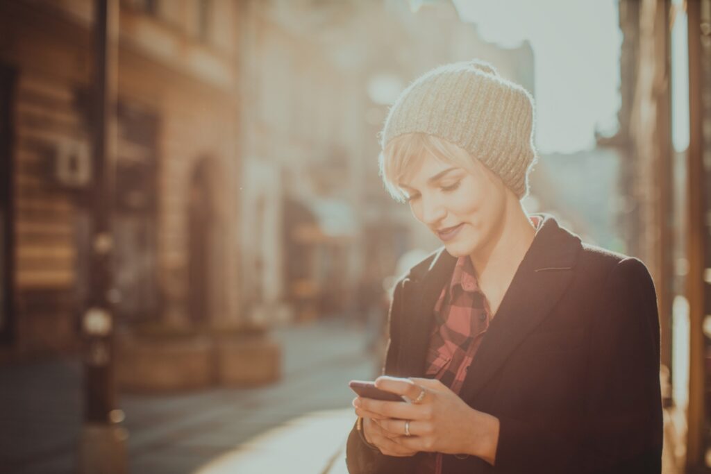 mulher envia mensagem pelo celular em dia de sol, com roupas de frio
